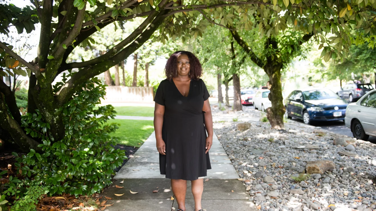 A Black woman stands under a tree and smiles, framed by green foliage all around. She is dressed in all black, with a dress and sandals and a silicone covered prosthetic leg.